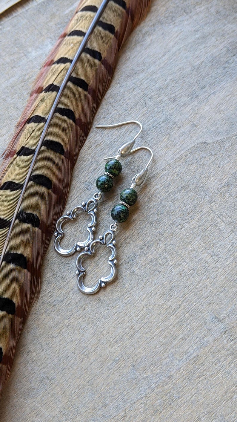 May include: Silver dangle earrings with two green and black speckled beads on each earring. The earrings have a floral design.