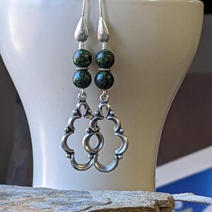 May include: Silver dangle earrings with two green and black speckled beads on each earring. The earrings have a decorative, openwork design.