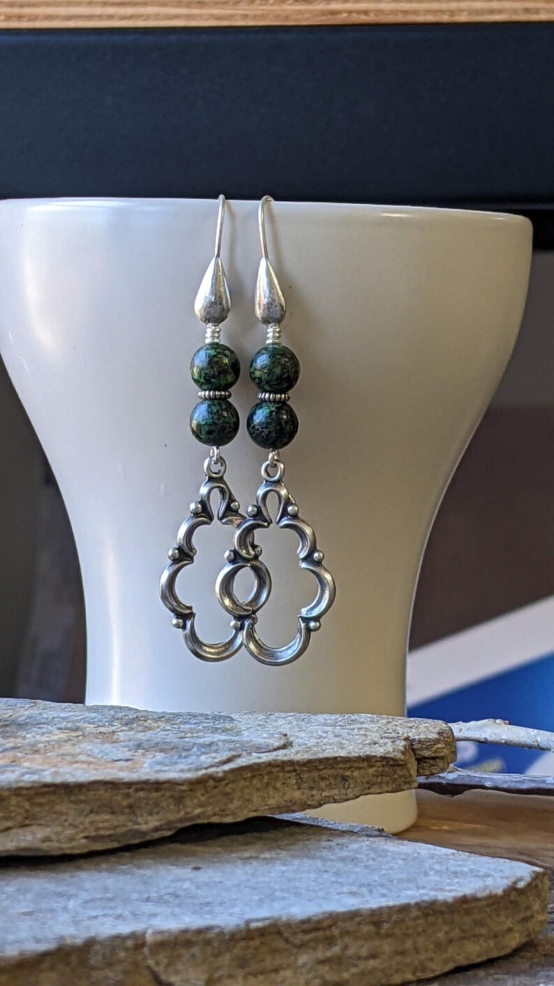 May include: Silver dangle earrings with two green and black speckled beads on each earring. The earrings have a decorative, openwork design.