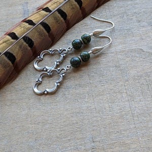 May include: Silver dangle earrings with green and black speckled stone beads. The earrings feature a delicate, ornate design.
