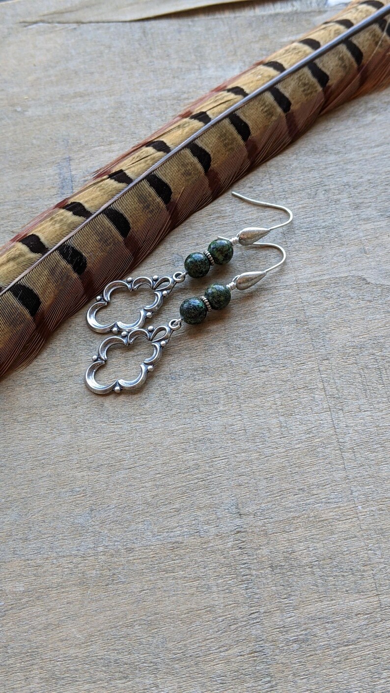 May include: Silver dangle earrings with green and black speckled stone beads. The earrings feature a delicate, ornate design.