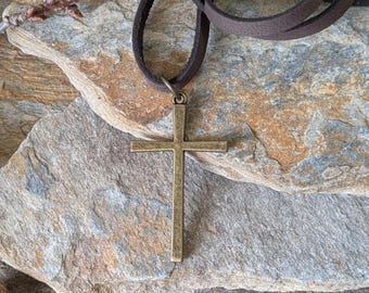 May include: A bronze-colored cross pendant necklace with a dark brown leather cord. The cross is a simple, classic design. The necklace is displayed on a textured, multi-toned stone surface.