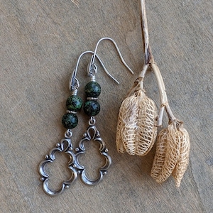 May include: Silver dangle earrings with a green and black speckled stone. The earrings have a filigree design.