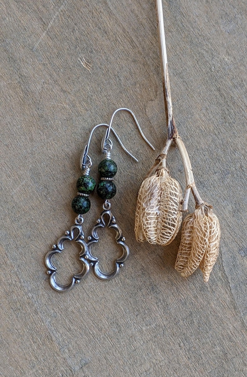 May include: Silver dangle earrings with a green and black speckled stone. The earrings have a filigree design.