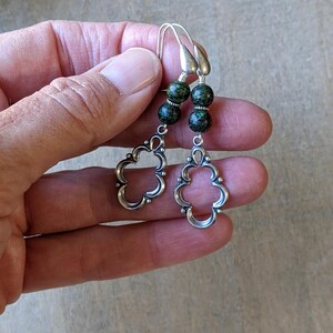 May include: Silver dangle earrings with green and gold speckled beads. The earrings feature a delicate, openwork design.
