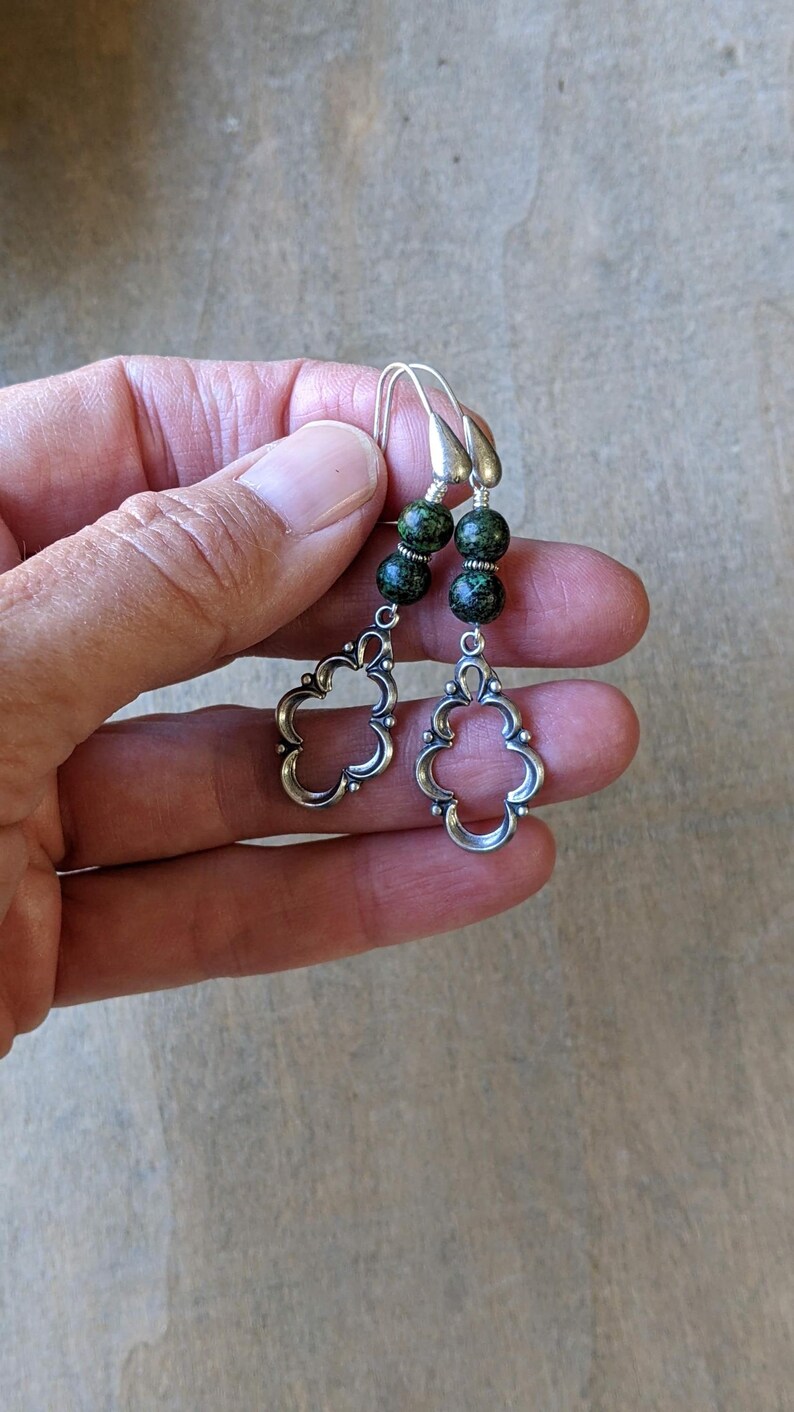 May include: Silver dangle earrings with green and gold speckled beads. The earrings feature a delicate, openwork design.