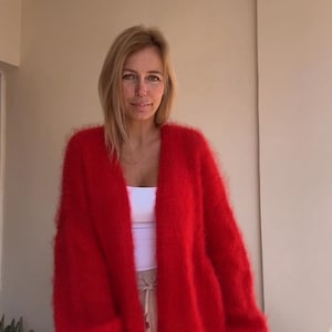 Puede incluir: Un cárdigan rojo brillante con frente abierto. El tejido de punto suave y esponjoso tiene mangas largas y un ajuste relajado. El cárdigan se usa sobre una camiseta blanca y pantalones beige, creando un look en capas. El estilo general es casual y cómodo.