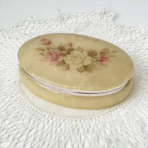 May include: Oval-shaped jewelry box with a cream-colored exterior and a silver-colored metal band. The lid features a floral design with pink and white roses and green leaves. The box is resting on a white, lacy surface.