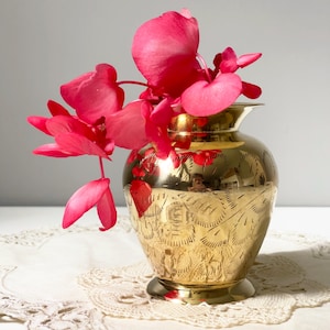 Peut inclure: Un vase en laiton doré, orné de motifs gravés, contient des fleurs roses vives. Le vase, de forme arrondie, présente un bord évasé. Les fleurs contrastent avec l'éclat métallique du vase.