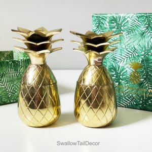 May include: Two gold pineapple shaped cocktail glasses. The glasses are made of metal and have a diamond pattern on the surface. The glasses are sitting on a white surface.