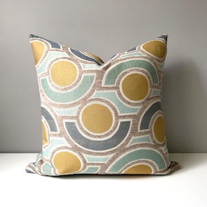 May include: Square decorative pillow with a geometric pattern. The design features overlapping circles and semi-circles in shades of gold, teal, gray, and tan. The pillow is made of a textured fabric and is likely used for home decor.