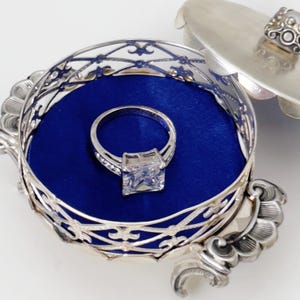 Op de afbeelding: Een zilveren ring met een grote, vierkante, heldere edelsteen ligt in een blauw fluwelen, sierlijke zilveren ringdoos. De doos is open en toont de ring. De doos heeft een decoratief filigraanontwerp en een bijpassend deksel.