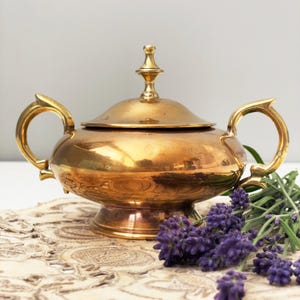May include: A vintage brass sugar bowl with a lid and two handles. The bowl has a pedestal base and a decorative finial on the lid. The handles have a unique, curved design. The bowl is displayed with sprigs of purple lavender.