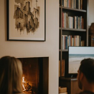 May include: A framed watercolor painting of a cityscape in shades of gray and brown hangs on a cream-colored wall. A fireplace with a burning fire is below the painting. A bookshelf filled with books is in the background.