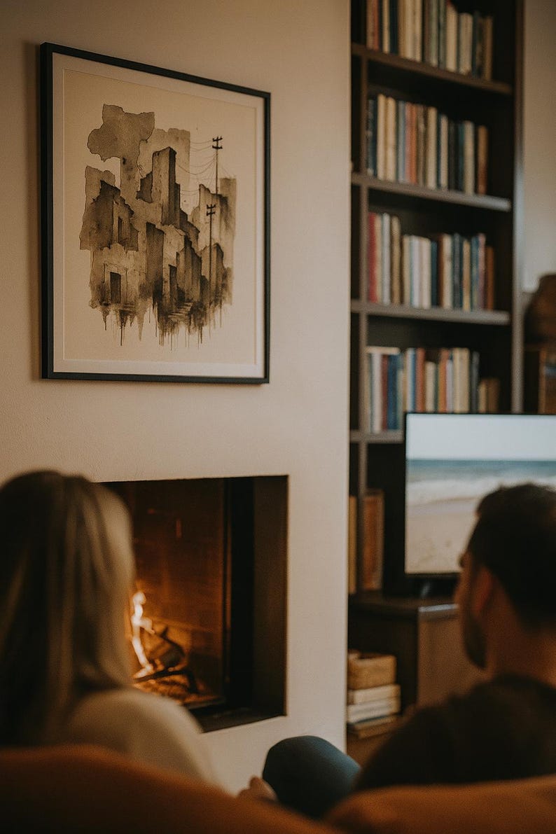 May include: A framed watercolor painting of a cityscape in shades of gray and brown hangs on a cream-colored wall. A fireplace with a burning fire is below the painting. A bookshelf filled with books is in the background.