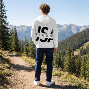 Puede incluir: Chaqueta con capucha blanca con el texto "USA FREEDOM" en negro, vista desde atrás. La chaqueta se usa con vaqueros azules y zapatillas blancas. El fondo presenta una cordillera y un cielo azul claro.
