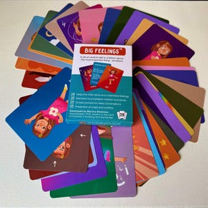 May include: A deck of "BIG FEELINGS" cards for children, featuring colorful illustrations and text. The box and cards are arranged in a circular pattern. The cards are designed to help children understand and talk about emotions. The box includes the number 39.