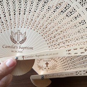 May include: Wooden hand fans with intricate cut-out designs. Each fan features a cross, doves, and laurel wreath, with the text "Camila's baptism 06.26.2026" printed in brown. Ideal for baptism favors or religious events.