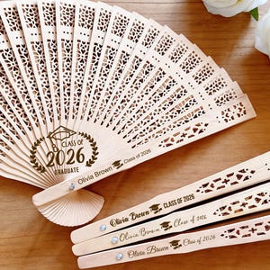May include: A collection of light-colored wooden fans, some open and some closed. The open fan displays the text "Class of 2026 Graduate" and the name "Olivia Brown". The fan blades have a decorative cut-out design. Additional fans are visible.
