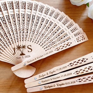 May include: A light-colored wooden fan with intricate cut-out designs. The fan is personalized with the initials "J S", palm trees, and the date "Nov 5, 2026". Additional fans are engraved with names and dates, perfect for a wedding or special event.