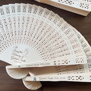 May include: A collection of intricately carved wooden fans. Each fan features a delicate openwork design and the inscription "In Loving Memory of Gabriel Smith 1956-2026" with a dove illustration. The fans are light in color.