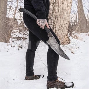 May include: A large, curved blade with a dark, mottled finish and a textured black handle. The knife features a prominent, pointed tip and a wide, sweeping edge. The metal surface displays a distinct, irregular pattern across the entire blade.