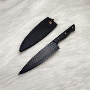 May include: A black chef's knife with a textured blade and a matching black handle. The knife is accompanied by a black leather sheath with a button closure. The blade has a unique hammered pattern.