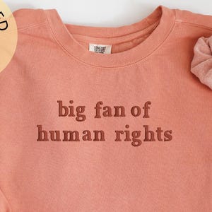 May include: Terracotta-colored sweatshirt with the text "big fan of human rights" in a darker shade. A matching scrunchie rests on the sleeve. The image also includes a circular logo that says "embroidered".