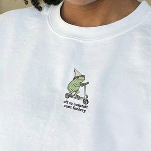 May include: White sweatshirt with a small graphic of a frog on a scooter wearing a wizard hat. The text below the graphic reads "off to commit some foolery". The sweatshirt is a crew neck style.