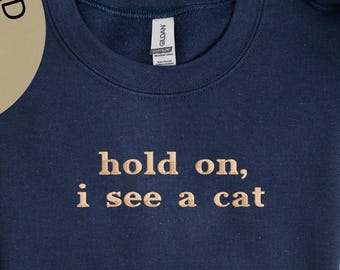 May include: Navy blue sweatshirt with the phrase "hold on, i see a cat" in a light brown font. The word "EMBROIDERED" is above a needle and thread graphic. The word "NAVY" is in the bottom right corner.