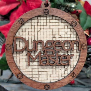 May include: A circular wooden ornament featuring the text Dungeon Master in raised dark wood letters over a light wood grid background. The outer frame is engraved with various polyhedral dice shapes at regular intervals around the perimeter.