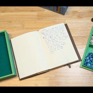 May include: An open, brown leather-bound journal with lined pages and a ribbon bookmark. Handwritten notes fill the right page. A green box and dice are visible on the right, suggesting a tabletop gaming theme.