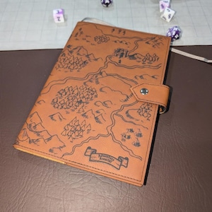 May include: A tan notebook cover featuring a black line-art map design with mountains, forests, and a banner with the text "Adventure Awaits". The cover includes a side flap with a silver-toned metal snap closure and visible stitching along the edges.
