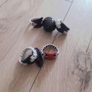 Peut inclure: Plusieurs bagues faites à la main avec des designs uniques. Les bagues présentent des accents noirs, blancs et rouges, certaines ayant un centre rouge. Une bague a une sphère noire. Les bagues sont disposées sur une surface en bois.