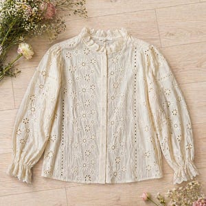 Op de afbeelding: Crèmekleurige blouse met broderie anglaise, een hoge, gerimpelde kraag en lange mouwen. De blouse heeft een knoopsluiting aan de voorkant en bloemvormige uitsparingen. Een boeket bloemen staat op de achtergrond.