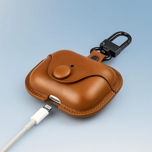 May include: A brown leather AirPod case with a black metal carabiner clip. The case has a button closure and a charging port. A white charging cable is plugged into the case. The background is a gradient of blue.