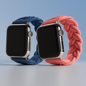 Pode incluir: Dois smartwatches com ecrãs pretos e caixas prateadas. Um tem uma pulseira trançada azul, o outro uma pulseira trançada coral. As pulseiras são feitas de um material espesso e tecido. O fundo é um gradiente azul claro.