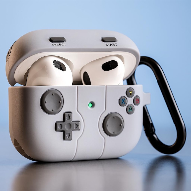 May include: A white and grey protective case for wireless earbuds, designed to look like a video game controller. The case has a directional pad, buttons, and the words "SELECT" and "START". A black carabiner clip is attached.