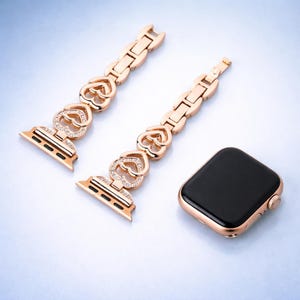 May include: Rose gold Apple Watch with a black screen and a matching rose gold band. The band features heart-shaped links with embedded crystals. The watch and band are displayed on a light blue surface.