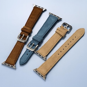 Puede incluir: Cuatro correas de reloj de ante en marrón, azul y beige. Cada correa tiene una hebilla y conectores plateados. Las correas están dispuestas en diagonal sobre un fondo azul claro, mostrando sus variaciones de textura y color.