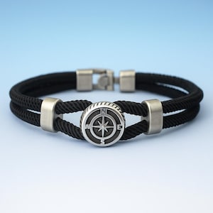 Puede incluir: Una pulsera de cuerda negra con un diseño de brújula plateada. La pulsera presenta un diseño de cuerda negra de doble hebra con detalles metálicos plateados y un cierre. El diseño de la brújula está centrado en la pulsera.