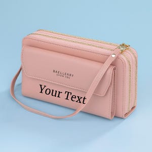May include: A light pink wallet-style crossbody bag with gold-colored zippers and hardware. The bag has a front flap pocket and a long, thin strap. The text "BAELLERY SHOW YOU" and "Your Text" are printed on the front.