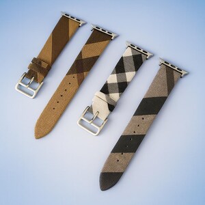 May include: Four patterned watch bands in shades of brown, tan, black, and white are displayed against a light blue background. Each band has a silver-colored buckle and connector. The patterns include stripes and a plaid design.