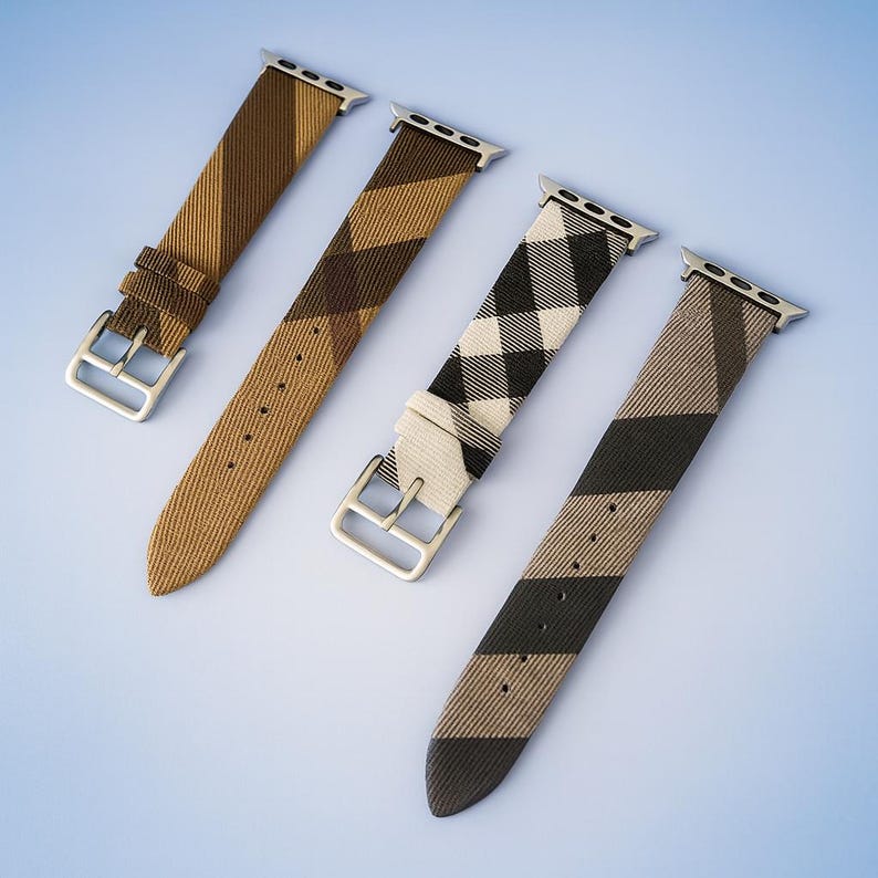 May include: Four patterned watch bands in shades of brown, tan, black, and white are displayed against a light blue background. Each band has a silver-colored buckle and connector. The patterns include stripes and a plaid design.