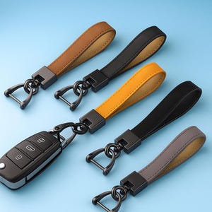 May include: A collection of leather keychains in various colors, including brown, black, yellow, and gray. Each keychain features a metal clasp and a loop for attaching keys. One keychain is attached to a black car key.