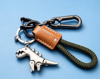 Könnte beinhalten: Ein Schlüsselanhänger mit einem silberfarbenen Metalldinosaurier-Anhänger, einer hellbraunen Lederlasche mit der Prägung HICRAFT und einer Schlaufe aus dunkelolivgrünem geflochtenem Seil. Das Set enthält einen schwarzen Karabiner und einen silbernen Ring.