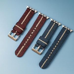 Puede incluir: Cuatro correas de reloj en tonos rojos y azules con una franja crema en el centro. Las correas tienen conectores plateados y hebillas doradas. Están dispuestas en diagonal sobre un fondo azul claro.