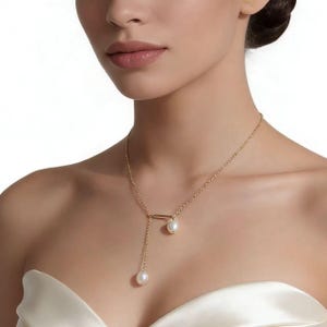 Puede incluir: Un collar dorado con un colgante de barra y dos perlas colgantes. El collar se lleva en el cuello y el pecho de una persona. Las perlas son blancas y redondas, y la cadena es delicada.