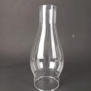 May include: A clear glass lamp chimney with a bulbous base that tapers into a narrow, cylindrical top. The glass is transparent and smooth, featuring a wide circular opening at the bottom and a smaller circular opening at the top for ventilation.
