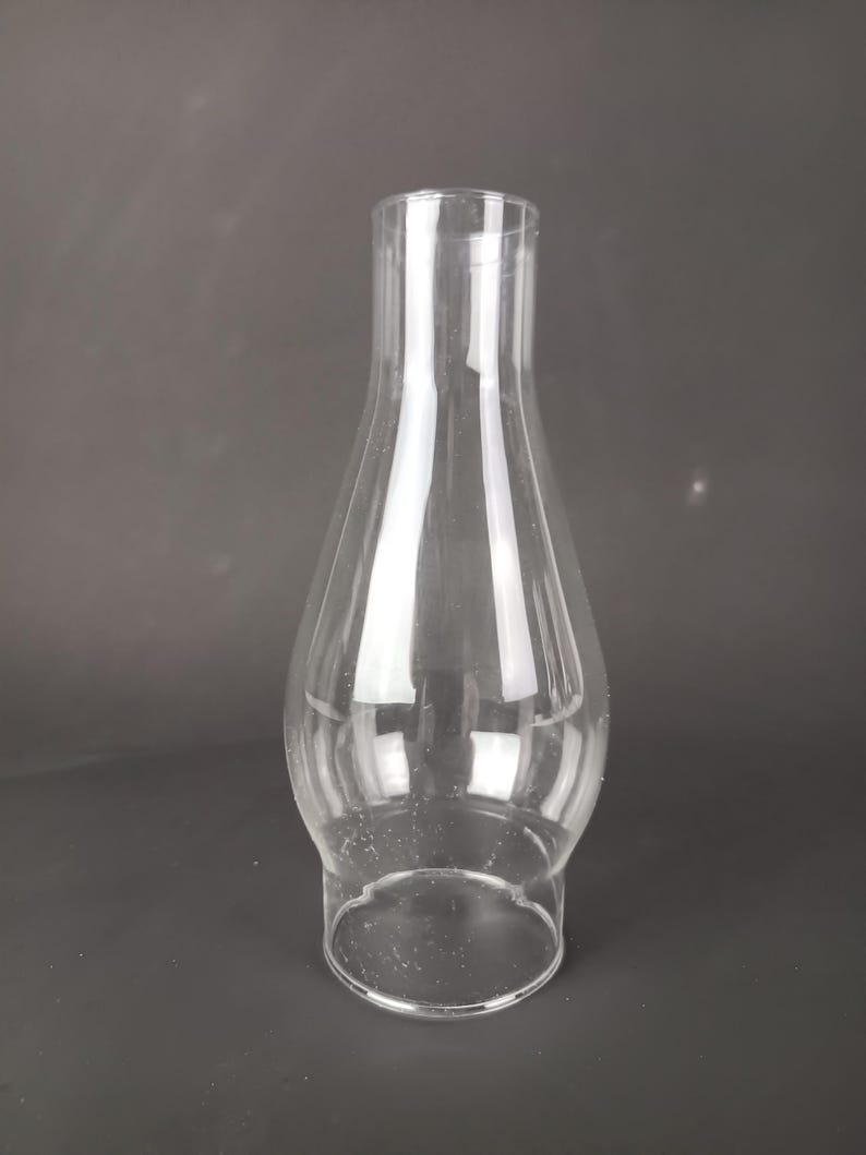May include: A clear glass lamp chimney with a bulbous base that tapers into a narrow, cylindrical top. The glass is transparent and smooth, featuring a wide circular opening at the bottom and a smaller circular opening at the top for ventilation.